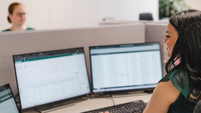 Back/profile view of female employee working, with a glimpse of a female employee across from her behind the screens and a desk divider