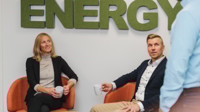 Employees Minna Lindholm and Arto Peltola sitting down in front of sign on the wall reading "energy" and having a chat with an employee entering the frame from the right