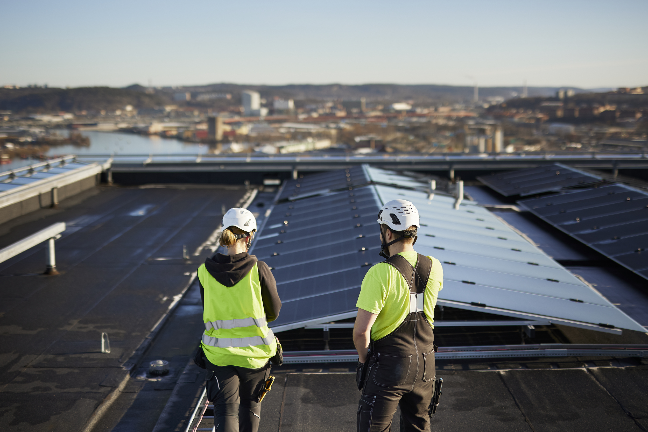 Two workers with bright yellow safety vests and helmets seen from behind looking at solar panels installed on the roof of a building