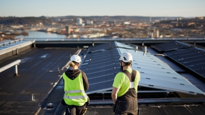 Two workers with bright yellow safety vests and helmets seen from behind looking at solar panels installed on the roof of a building