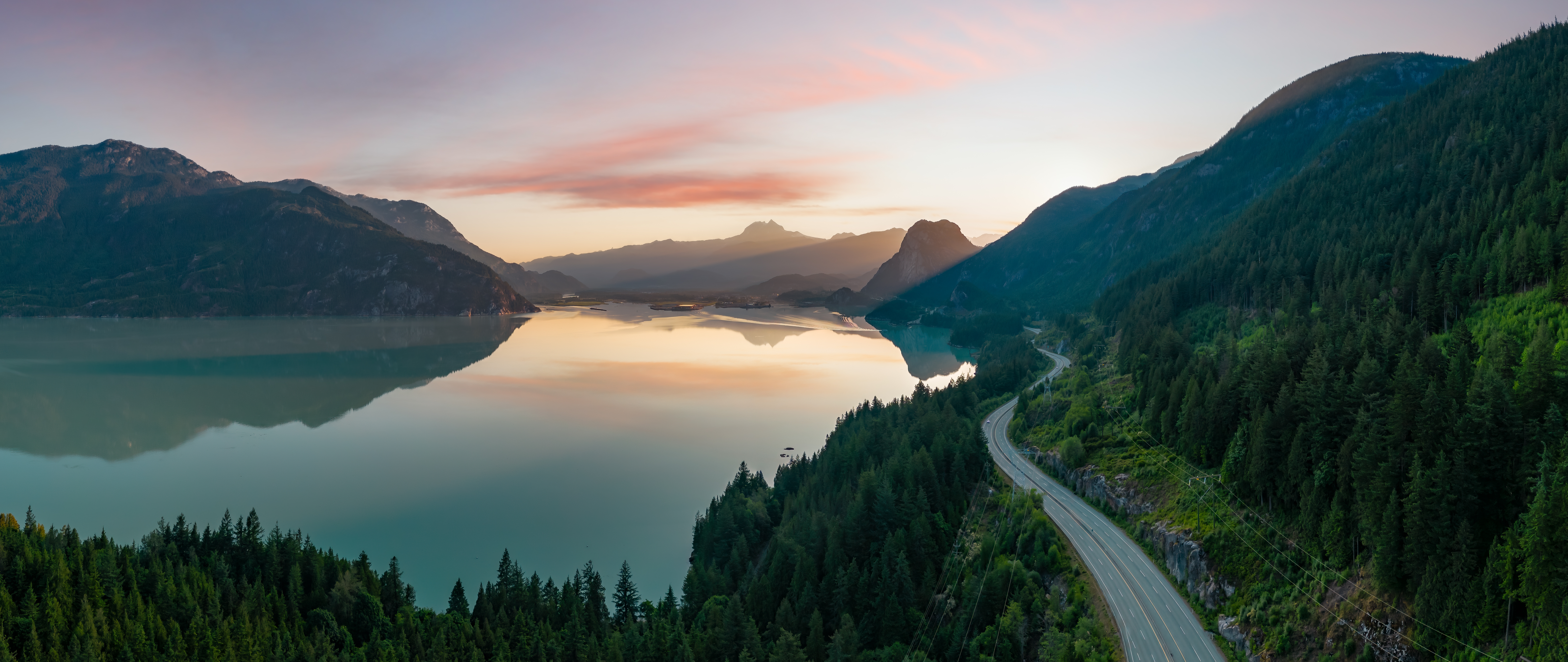 A scenic nature photo with a road running along mountains on the right side, and a big lake and mountains in the distance on the left